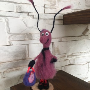 May include: A whimsical felt figurine of a purple creature with long black antennae, wearing black boots and carrying a purple bag. The figurine is standing on a wooden base.