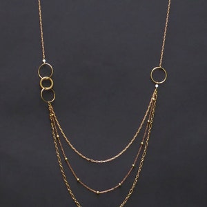Peut inclure: Un collier doré à plusieurs niveaux avec trois rangées de chaînes de styles différents. Le collier comporte de petites perles dorées et trois anneaux connectés d'un côté, et un seul grand anneau de l'autre. Le fond est gris foncé.