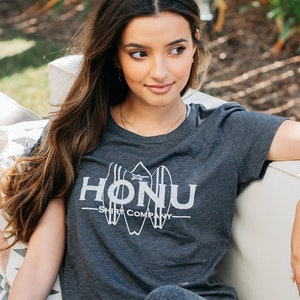 May include: A woman wearing a dark grey t-shirt with the text "HONU Surf Company" in white. She is sitting on a white couch with a patterned pillow behind her. She is wearing dark grey leggings and white sneakers.