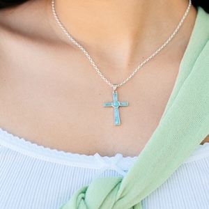 May include: A woman wearing a white tank top and a green scarf is wearing a silver necklace with a blue and silver cross pendant.