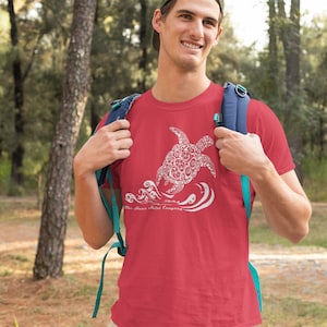 Puede incluir: Una camiseta roja con un gr&aacute;fico blanco de una tortuga marina estilizada nadando en olas. El texto "The Shore Shirt Company" est&aacute; impreso debajo de la tortuga.