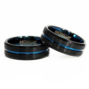 K&ouml;nnte beinhalten: Zwei schwarze Ringe aus Wolframcarbid mit einem blauen Streifen am Rand. Die Ringe sind mit dem Wort "Honux" graviert.