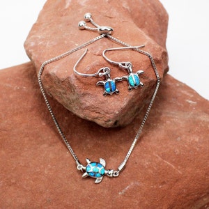 May include: A silver chain bracelet and matching earrings with a blue opal turtle charm. The jewelry is displayed on a red rock.