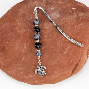Peut inclure: Signet en argent avec un charme de tortue en argent et une chaîne de perles avec des pierres noires et grises.