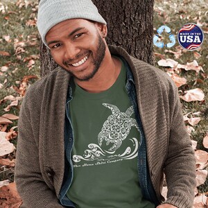 Puede incluir: Un hombre lleva una camiseta verde con un gr&aacute;fico blanco de una tortuga nadando en olas. La camiseta tiene el texto "The Aloha Shirt Company" impreso en la parte inferior. El hombre est&aacute; sentado en el suelo con un libro en su regazo.