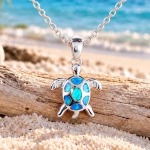 May include: A silver-toned necklace featuring a sea turtle pendant with a shell inlaid with iridescent blue and green opal-like stones. The pendant hangs from a delicate silver chain, showcasing a detailed turtle design with four flippers.