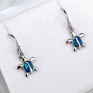 May include: A pair of silver turtle earrings with blue opal accents. The earrings are displayed in a white box with a teal lid.