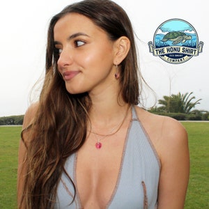 Op de afbeelding: Een vrouw draagt een lichtblauw haltertop met een zilveren ketting met een roze hart hanger. Ze draagt ook zilveren oorbellen met roze hart bedels. De vrouw heeft lang bruin haar en kijkt naar de zijkant.