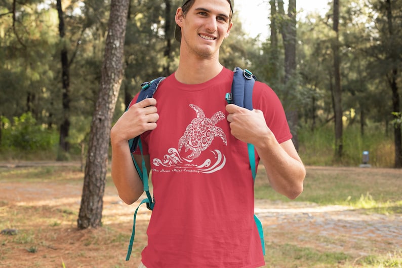 Puede incluir: Una camiseta roja con un gr&aacute;fico blanco de una tortuga marina estilizada nadando en olas. El texto "The Shore Shirt Company" est&aacute; impreso debajo de la tortuga.