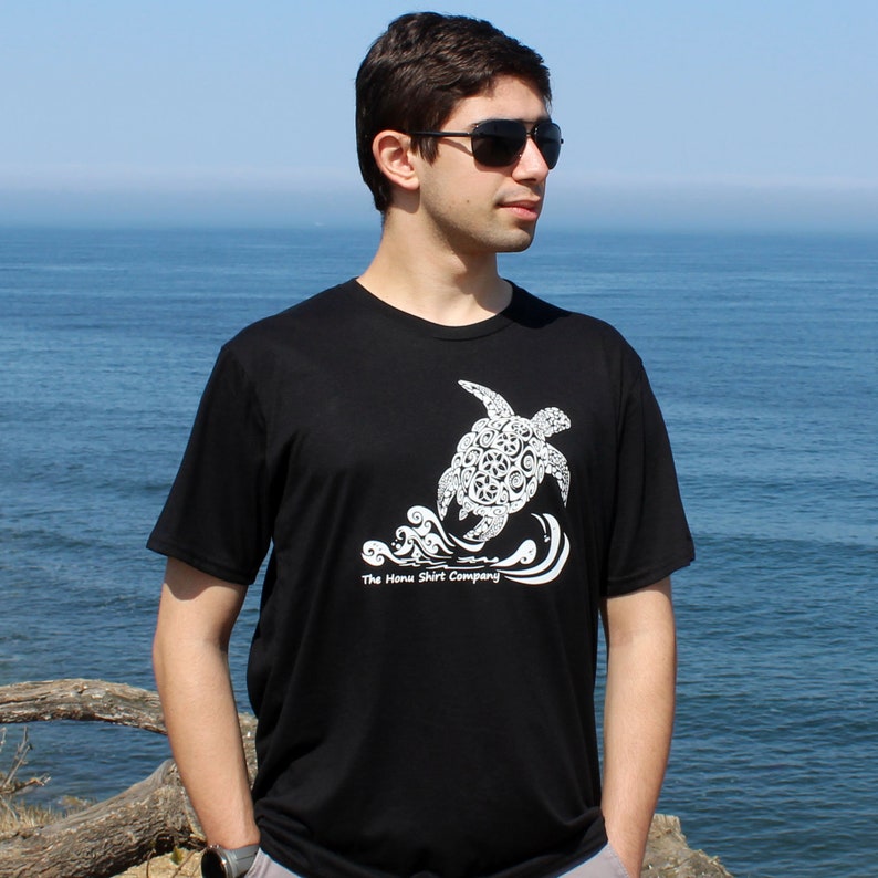 Puede incluir: Una camiseta negra con un gr&aacute;fico blanco de una tortuga marina estilizada nadando en olas. El texto "The Honu Shirt Company" est&aacute; impreso debajo de la tortuga.