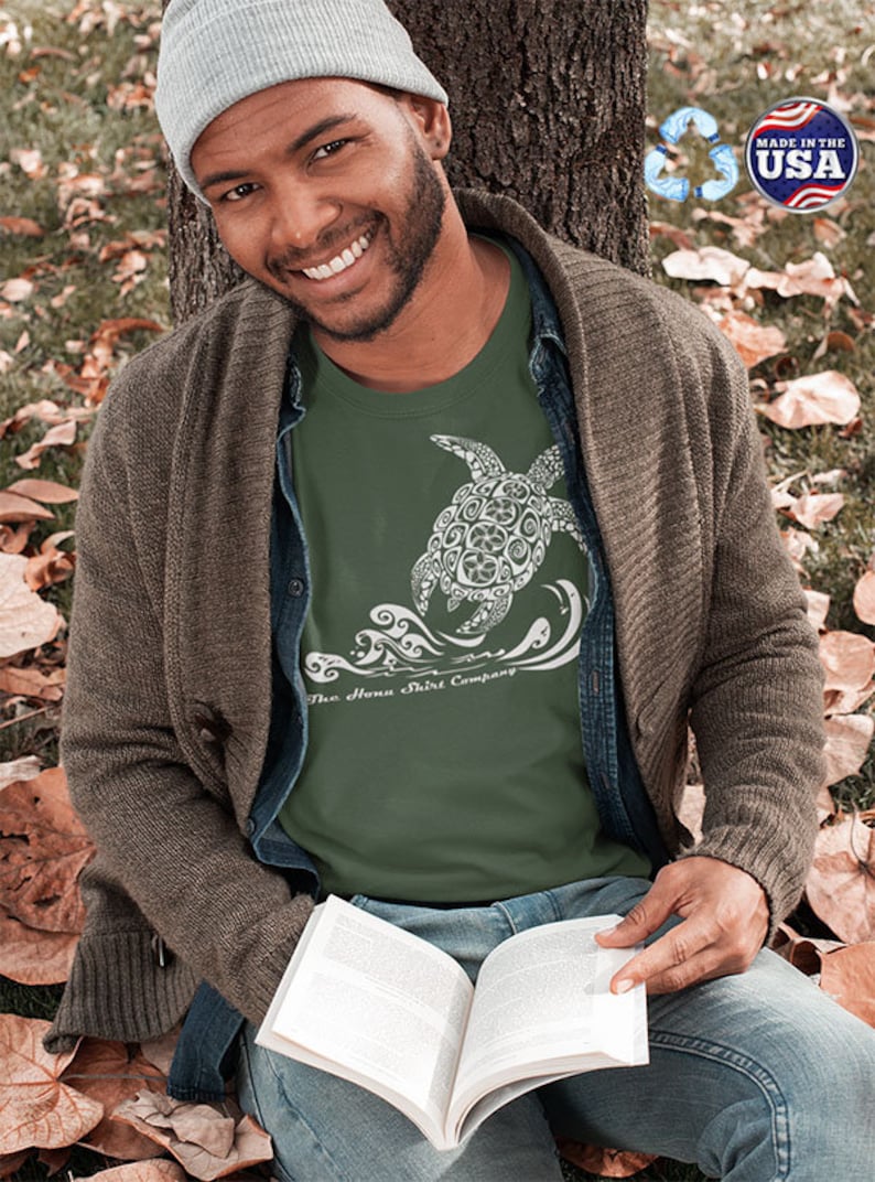Puede incluir: Un hombre lleva una camiseta verde con un gr&aacute;fico blanco de una tortuga nadando en olas. La camiseta tiene el texto "The Aloha Shirt Company" impreso en la parte inferior. El hombre est&aacute; sentado en el suelo con un libro en su regazo.
