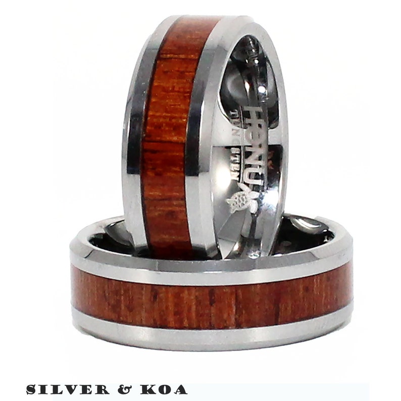 May include: Two silver and koa wood rings. The rings are stacked on top of each other. The top ring has the word "HONOLULU" engraved on the side. The bottom ring has a koa wood inlay.