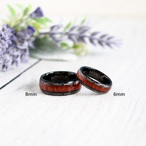 May include: Two black tungsten carbide rings with a wood inlay and silver engraving. The ring on the left is 8mm wide and the ring on the right is 6mm wide. Both rings have the text "Honu Tungsten" engraved on the inside.