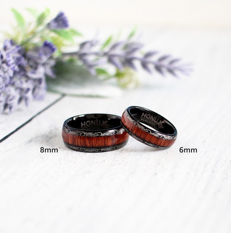 May include: Two black tungsten carbide rings with a wood inlay and silver engraving. The ring on the left is 8mm wide and the ring on the right is 6mm wide. Both rings have the text "Honu Tungsten" engraved on the inside.