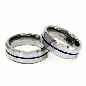 K&ouml;nnte beinhalten: Zwei silberne Ringe aus Wolframcarbid mit einer blauen Linien-Einlage. Die Ringe sind abgeschr&auml;gt und haben das Wort "HONOR" auf der Innenseite eingraviert.