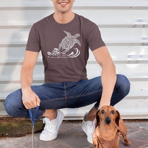 Puede incluir: Un hombre lleva una camiseta marr&oacute;n con un gr&aacute;fico blanco de una tortuga nadando en el oc&eacute;ano. La camiseta tiene el texto "The Ocean Tribe Company" impreso en ella. Est&aacute; en cuclillas y sostiene una correa atada a un dachshund marr&oacute;n.