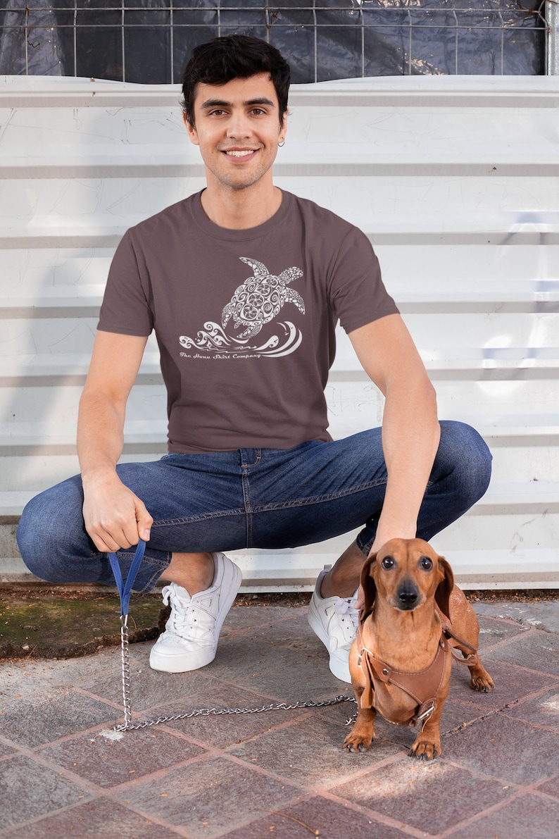 Puede incluir: Un hombre lleva una camiseta marr&oacute;n con un gr&aacute;fico blanco de una tortuga nadando en el oc&eacute;ano. La camiseta tiene el texto "The Ocean Tribe Company" impreso en ella. Est&aacute; en cuclillas y sostiene una correa atada a un dachshund marr&oacute;n.