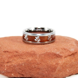 May include: A silver ring with a brown wood inlay and a design of sea turtles. The ring is sitting on a red rock.