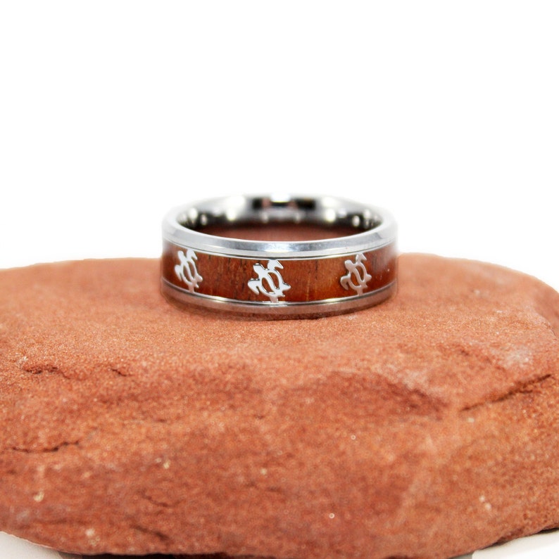 May include: A silver ring with a brown wood inlay and a design of sea turtles. The ring is sitting on a red rock.