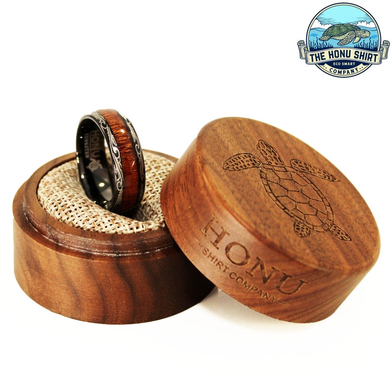 May include: A black and brown ring with a wood inlay and silver engraving is displayed in a wooden box with a turtle design and the text "HONU - SHIRT COMPANY".
