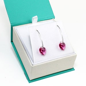 Op de afbeelding: Een paar zilveren hartvormige oorbellen met roze kristallen accenten, gepresenteerd in een turquoise en witte geschenkdoos.