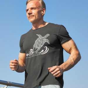 Puede incluir: Un hombre lleva una camiseta negra con un gr&aacute;fico blanco de una tortuga estilizada nadando en olas. El texto "The Ocean Shirt Company" est&aacute; impreso debajo de la tortuga.