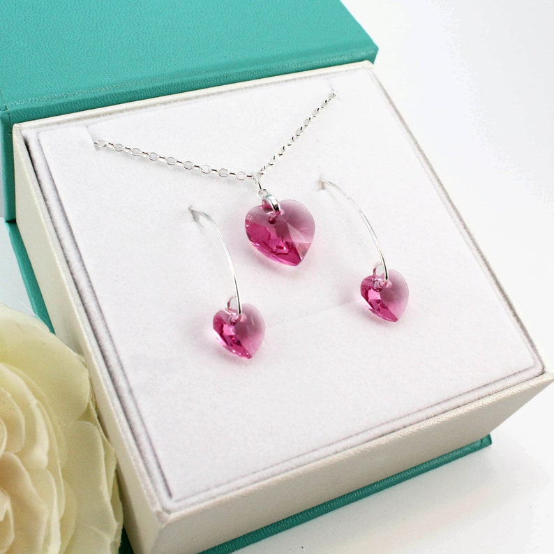 Op de afbeelding: Een roze kristallen hart ketting en oorbellen set in een witte doos. De ketting heeft een zilveren ketting en de oorbellen zijn zilveren hoepels met een roze kristallen hart dat aan elke hoepel hangt.