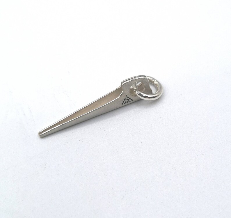 K&ouml;nnte beinhalten: Ein silberfarbener Metallanh&auml;nger in Form einer scharfen Spitze mit einem kleinen Ring oben. Der Anh&auml;nger hat ein Dreieck mit den Buchstaben "J.A.D." darin.