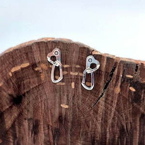Può includere: Un paio di orecchini d'argento a forma di moschettoni da arrampicata in miniatura. Gli orecchini sono esposti su un pezzo di legno.