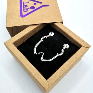 Puede incluir: Pendientes plateados en una caja de cartón marrón con un logotipo morado. Los pendientes tienen un diseño curvo único con pequeños detalles circulares. La caja está abierta, mostrando los pendientes sobre una superficie de terciopelo negro.