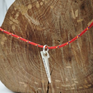 以下が含まれることがあります： 銀製のピトン型のペンダント。42.20.20.3' N 2.16.56.7' Eの座標が刻まれています。ペンダントは赤と青のロープにぶら下がっています。