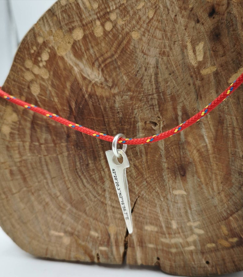 K&ouml;nnte beinhalten: Silberner Anh&auml;nger in Form eines Kletterhammers mit den Koordinaten 42.20.20.3' N 2.16.56.7' E eingraviert. Der Anh&auml;nger h&auml;ngt an einem rot-blauen Seil.