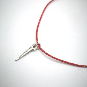 K&ouml;nnte beinhalten: Eine rote Schnurkette mit einem silbernen Anh&auml;nger in Form eines spitzen Objekts. Der Anh&auml;nger tr&auml;gt die Gravur "9.7.20.19.N.Z.T.E".