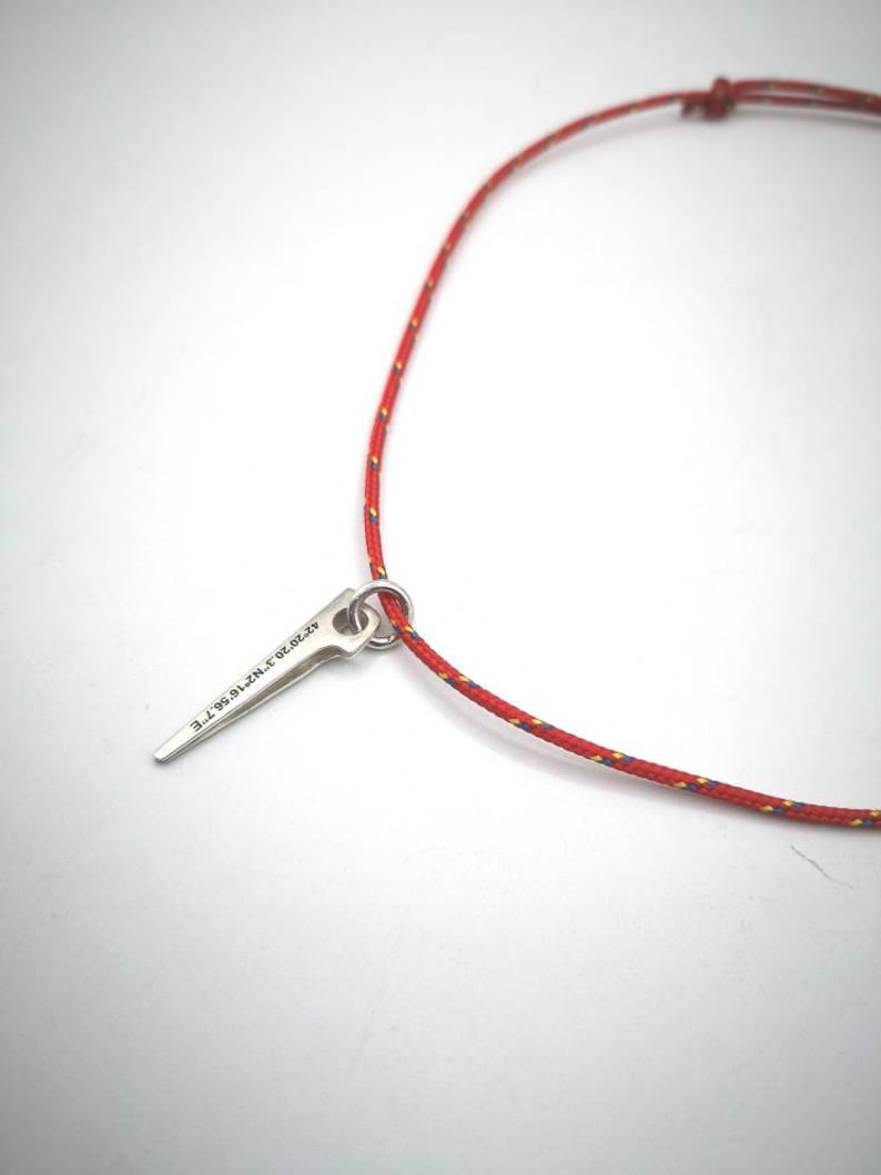 K&ouml;nnte beinhalten: Eine rote Schnurkette mit einem silbernen Anh&auml;nger in Form eines spitzen Objekts. Der Anh&auml;nger tr&auml;gt die Gravur "9.7.20.19.N.Z.T.E".