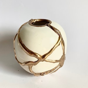 Puede incluir: Jarrón de cerámica blanco con un esmalte metálico dorado. El esmalte se aplica en un patrón de telaraña.