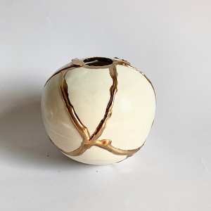 Könnte beinhalten: Weiße Keramikvase mit goldenem Metallic-Glanz-Design. Die Vase ist rund und hat eine kleine Öffnung oben.