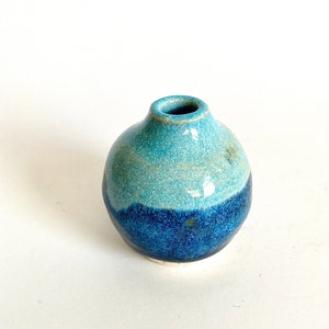 Puede incluir: Un pequeño jarrón redondo de cerámica con un esmalte azul y azul claro. El jarrón tiene una abertura ligeramente redondeada en la parte superior.