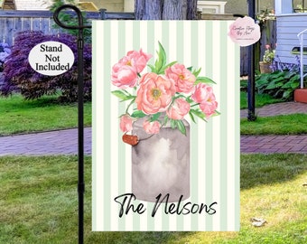 May include: A green and white striped garden flag with a watercolor illustration of pink peonies in a gray metal pitcher. The text "The Nelsons" is printed below the pitcher.