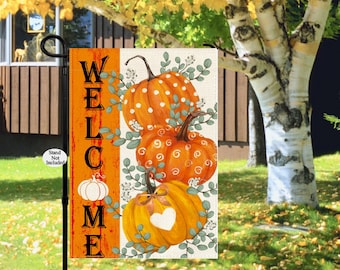 May include: A garden flag with a fall theme. The flag features three orange pumpkins with decorative designs and green foliage. The word "WELCOME" is printed vertically in black on an orange background. The flag is displayed on a black stand.