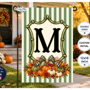 May include: A rectangular garden flag featuring a vertical light green and cream striped background. A central cream shield with a black letter M is framed by a green border. Below, a cluster of pumpkins, autumn leaves, and a plaid bow.