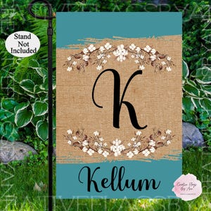 May include: A teal blue garden flag with a burlap background featuring a white cotton flower wreath with a black letter K in the center. The text "Kellum" is printed in black at the bottom of the flag.