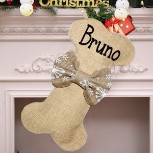 May include: A tan burlap Christmas stocking shaped like a dog bone. The name Bruno is printed in black script on the upper section. A matching burlap bow with a white snowflake pattern is attached to the center of the stocking.