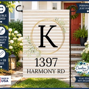 May include: A rectangular garden flag featuring a beige and white horizontal striped pattern. The center displays a large black letter K inside a gold circular frame with green leaf accents. Below, the text reads 1397 HARMONY RD in black.