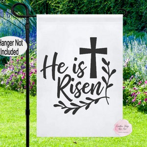 Pode incluir: Bandeira de jardim branca com texto preto e uma cruz. O texto diz "He is Risen" com um desenho floral abaixo da cruz.