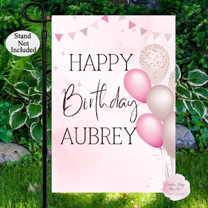 May include: Pink and gold Happy Birthday garden flag with the name Aubrey. The flag features pink and gold balloons with confetti and a banner of pink and gold triangles.