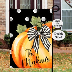 May include: A double-sided garden flag featuring a watercolor pumpkin with a black and white striped bow. The flag has a black background with white polka dots and the text "The Makua's" in a script font. The flag is designed for outdoor use.