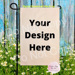 May include: A beige garden flag mockup with the text "Your Design Here" in black. The flag is hanging on a black metal pole in front of a blue wooden fence and green grass with white flowers.