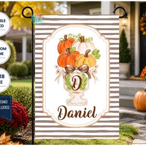 May include: A rectangular garden flag featuring a brown and white horizontal striped background. The center displays a white decorative frame containing a watercolor illustration of pumpkins in an urn, a brown bow, and the name Daniel in script.