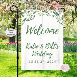 May include: A garden flag with a white wooden plank background and green text that reads "Welcome Katie & Bill's Wedding JUNE 29, 2024". The top border features green leaves and floral accents. A black metal stand is holding the flag.