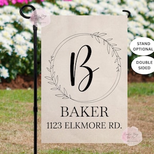 May include: A beige garden flag with a black script letter "B" inside a circular wreath design. The name "BAKER" and address "1123 ELKMORE RD." are printed below. The flag is double-sided.